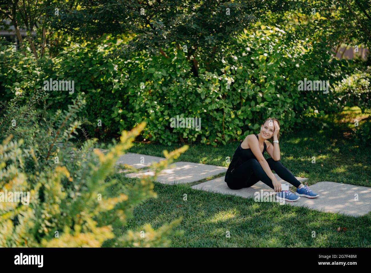 Prise de vue horizontale en longueur d'une femme sportive brune heureuse écoute la playlist de motivation avant de commencer à courir vêtue de poses de vêtements actifs sur la trajectoire Banque D'Images