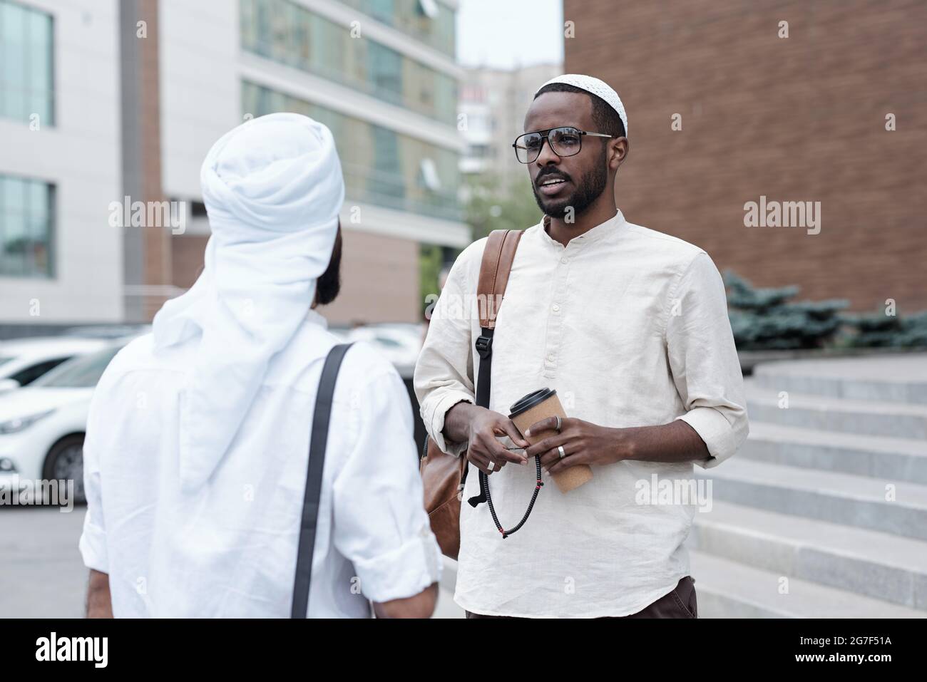 Un jeune musulman confiant dans des lunettes debout dans la rue et en train de boire un café avec un partenaire d'affaires Banque D'Images