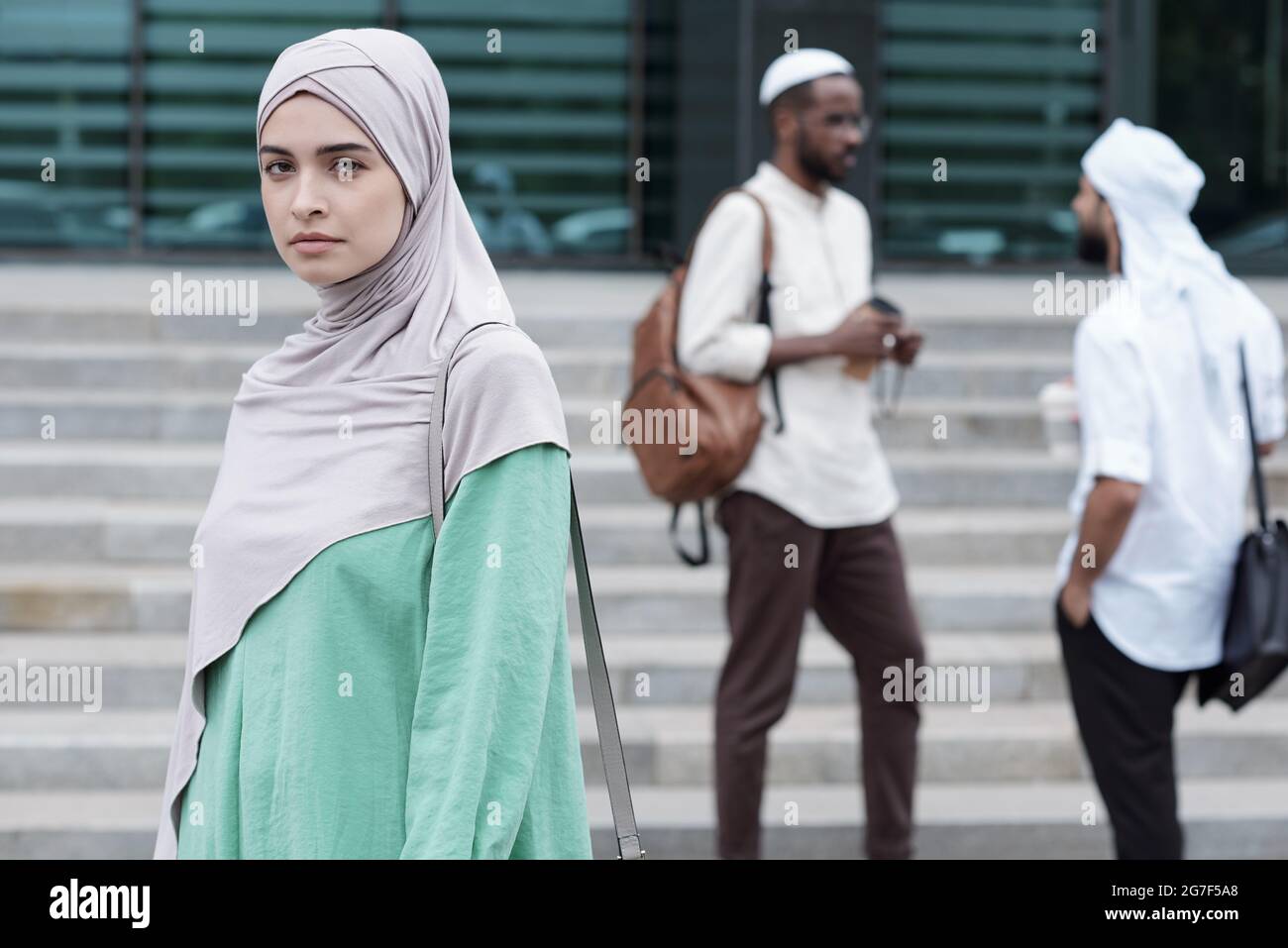 Portrait d'une jeune femme musulmane sérieuse et confiante en vêtements nationaux portant un sac à bandoulière contre ses collègues à l'extérieur Banque D'Images