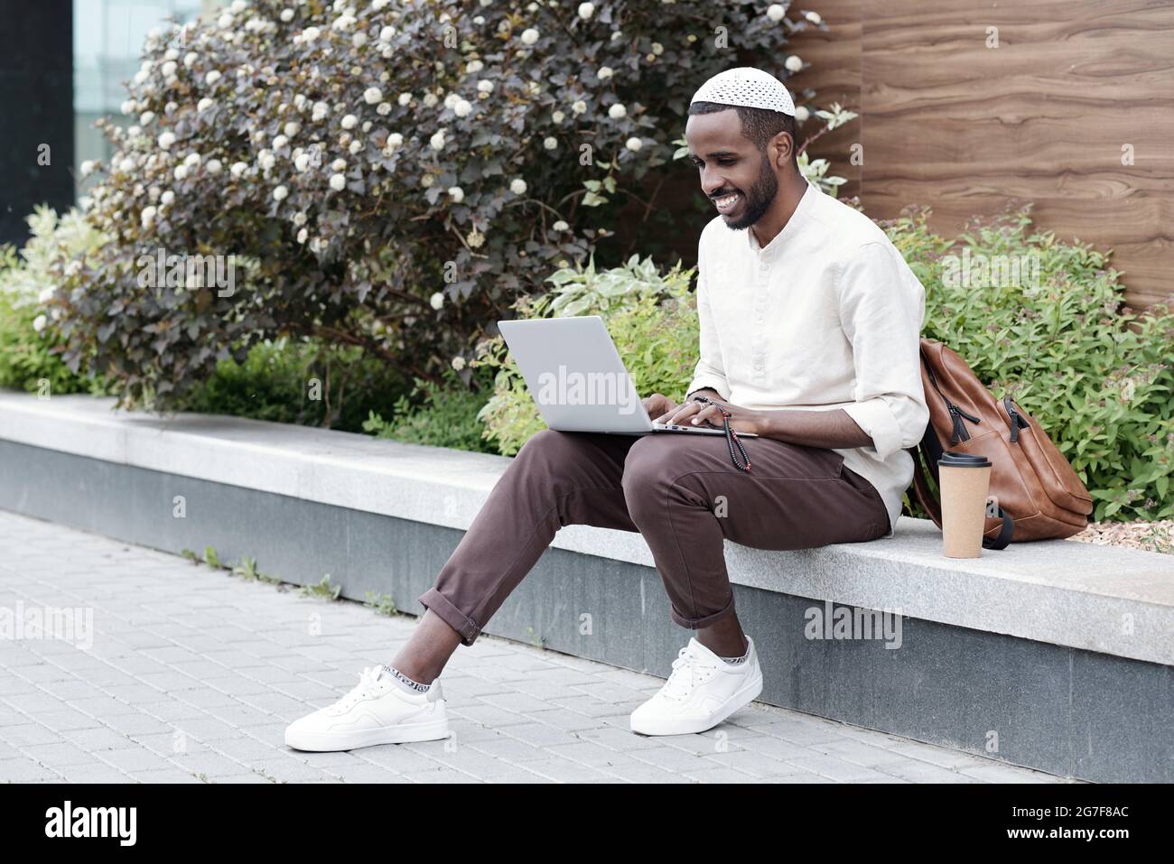 Un jeune acheteur musulman positif en casquette kufi assis sur le trottoir et utilisant un ordinateur portable tout en travaillant en ligne Banque D'Images