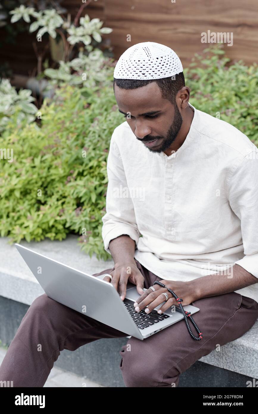 Concentré de jeunes musulmans occupés dans le chapeau kufi assis sur le trottoir et travaillant avec un ordinateur portable Banque D'Images