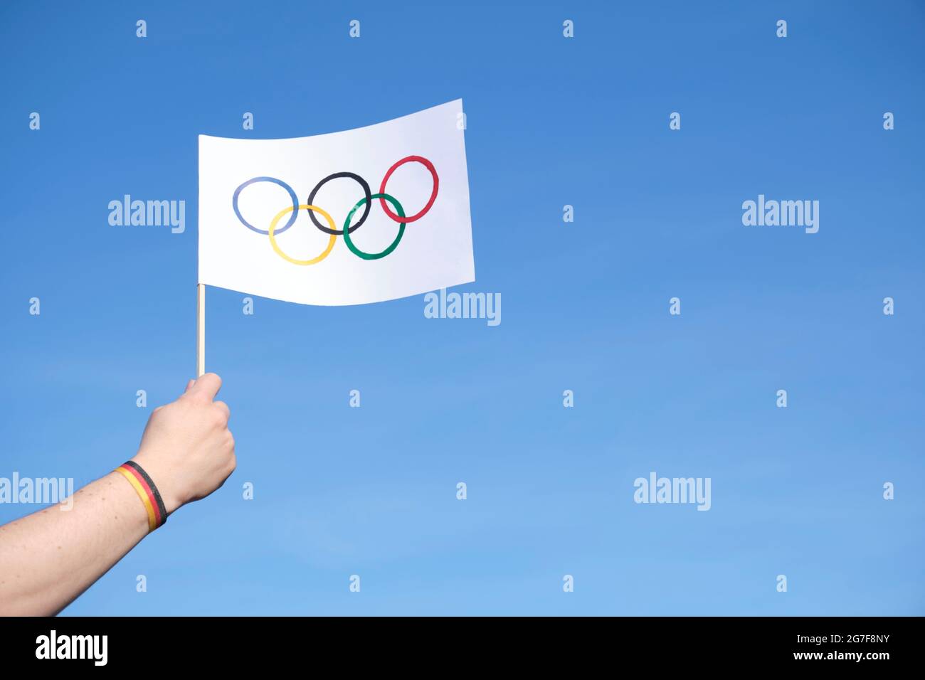 Main tenant un drapeau artisanal des Jeux Olympiques à l'extérieur, contre un ciel clair, portant un bracelet bleu, blanc et rouge, couleurs du drapeau national allemand. Banque D'Images