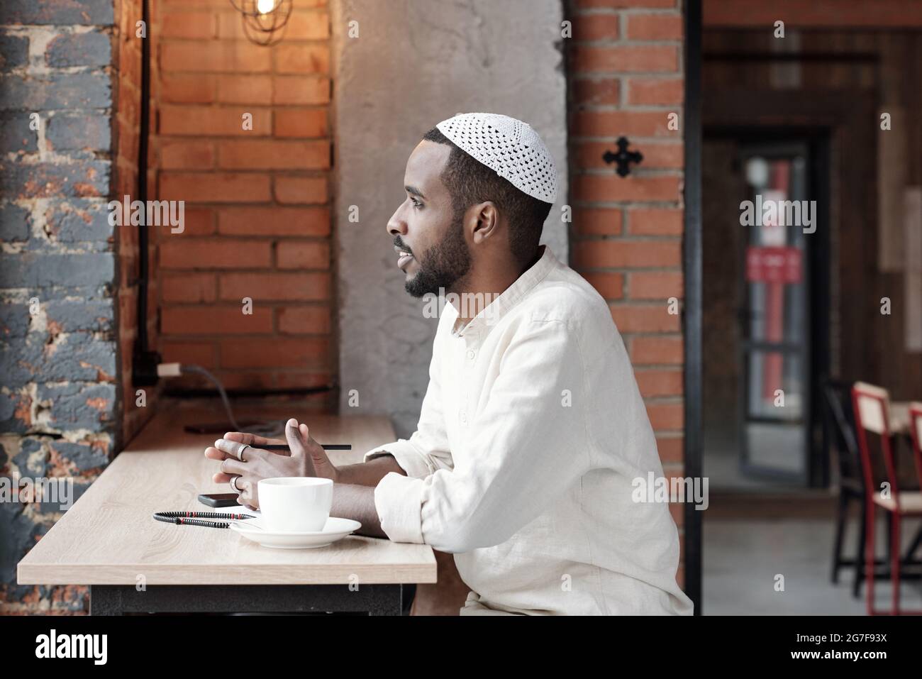 Jeune homme islamique pensif inspiré en casquette kufi assis à une table dans le café loft et donnant sur la fenêtre Banque D'Images