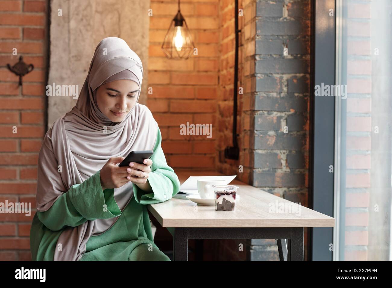 Contenu jeune femme musulmane dans hijab assis à une table haute dans le café loft et SMS sur le téléphone Banque D'Images