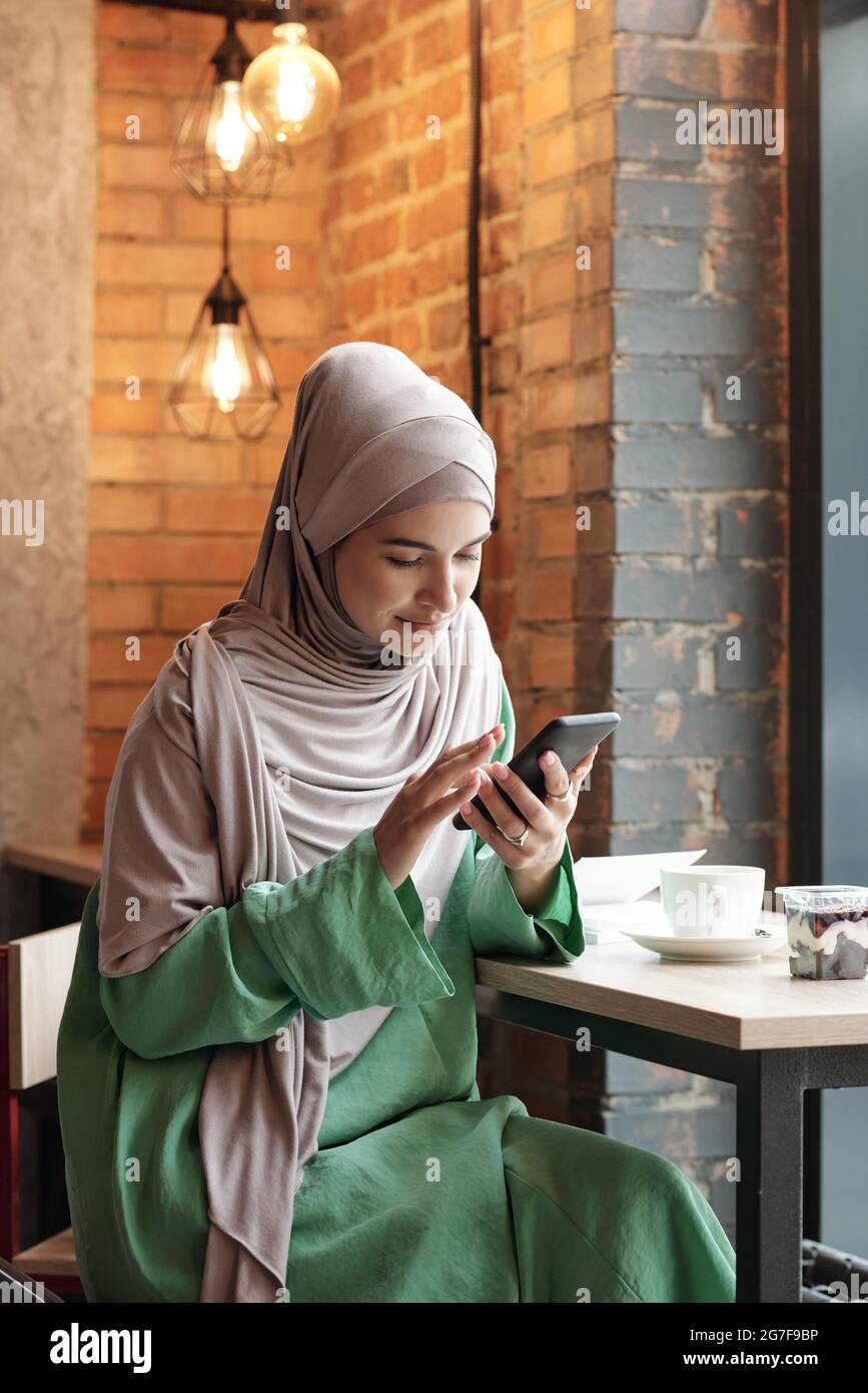 Contenu jeune femme musulmane dans hijab assis à table et boire un café tout en se connectant au WIFI dans le café Banque D'Images