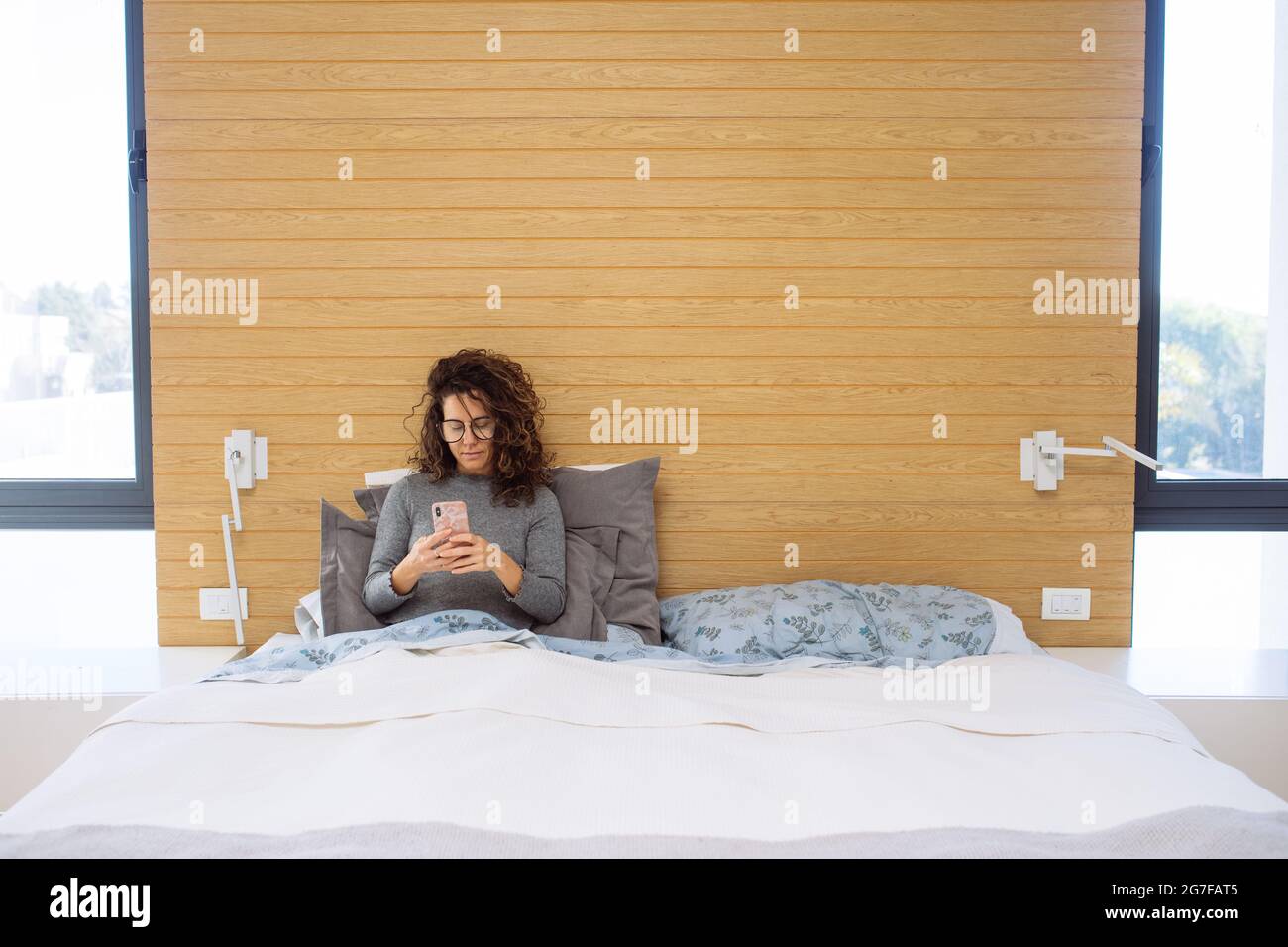 Femme de race blanche dans sa trentaine dans un lit double king size regardant et tapotant son smartphone sur un côté du lit. Envoyer des SMS à son mari. Banque D'Images