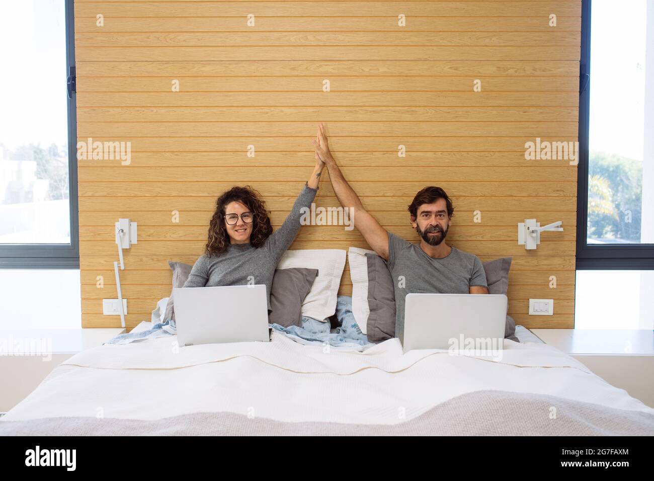 Couple dans un lit double king size chacun avec leur ordinateur portable faisant un cinq hi regardant l'appareil photo. Concept : succès du bureau à domicile Banque D'Images