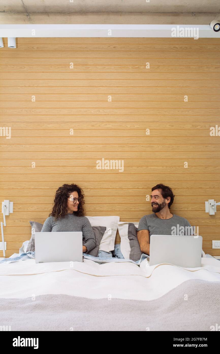 Couple dans un lit double king size chacun avec leur ordinateur portable de travail regardant l'un l'autre et souriant. Concept : bureau à domicile Banque D'Images