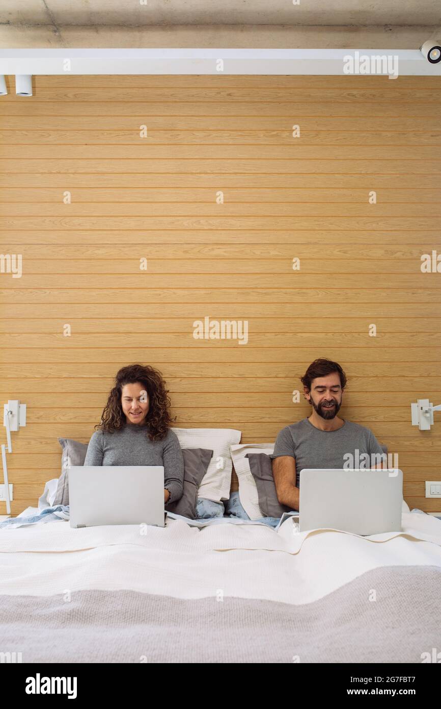 Couple dans un lit double king size chacun avec leur ordinateur portable de travail regardant chacun de leurs écrans et souriant. Concept : bureau à domicile Banque D'Images