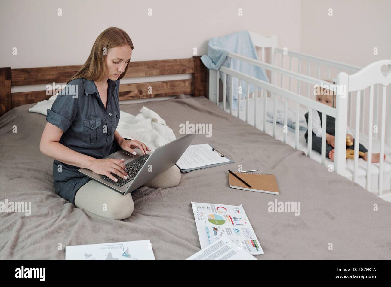 Jeune mère de bébé concentrée assise sur le lit et utilisant un ordinateur portable pendant que son fils la regarde de son berceau Banque D'Images