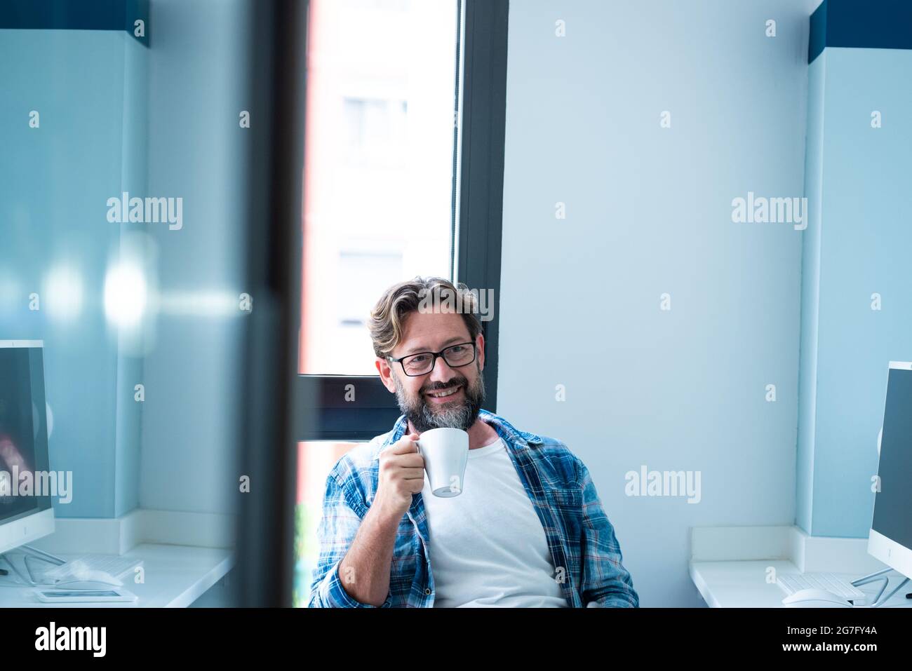 Joyeux homme barbu adulte assis au bureau sourire et profiter de pause de travail en ligne - les gens et le style de vie de travail - les couleurs bleues - beau mal mûr Banque D'Images