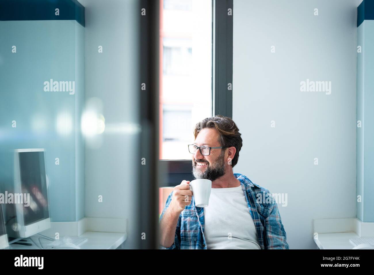 Portrait de l'homme heureux regardant à l'extérieur de la fenêtre et sourire en buvant un café à la maison ou au bureau - mâle caucasien adulte avec la barbe et les lunettes dans le travail br Banque D'Images
