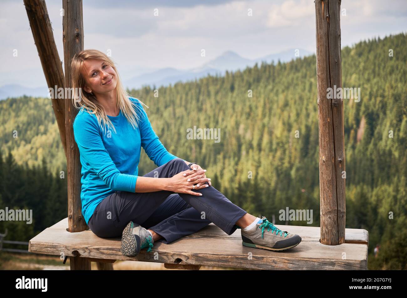 Portrait d'une gentille dame dans un chemisier bleu qui mignonne assis et souriant tout en se relaxant sur une énorme balançoire en bois à la chaude journée d'été. Sur la forêt de fond, silhouettes de collines de montagne et ciel gris. Banque D'Images