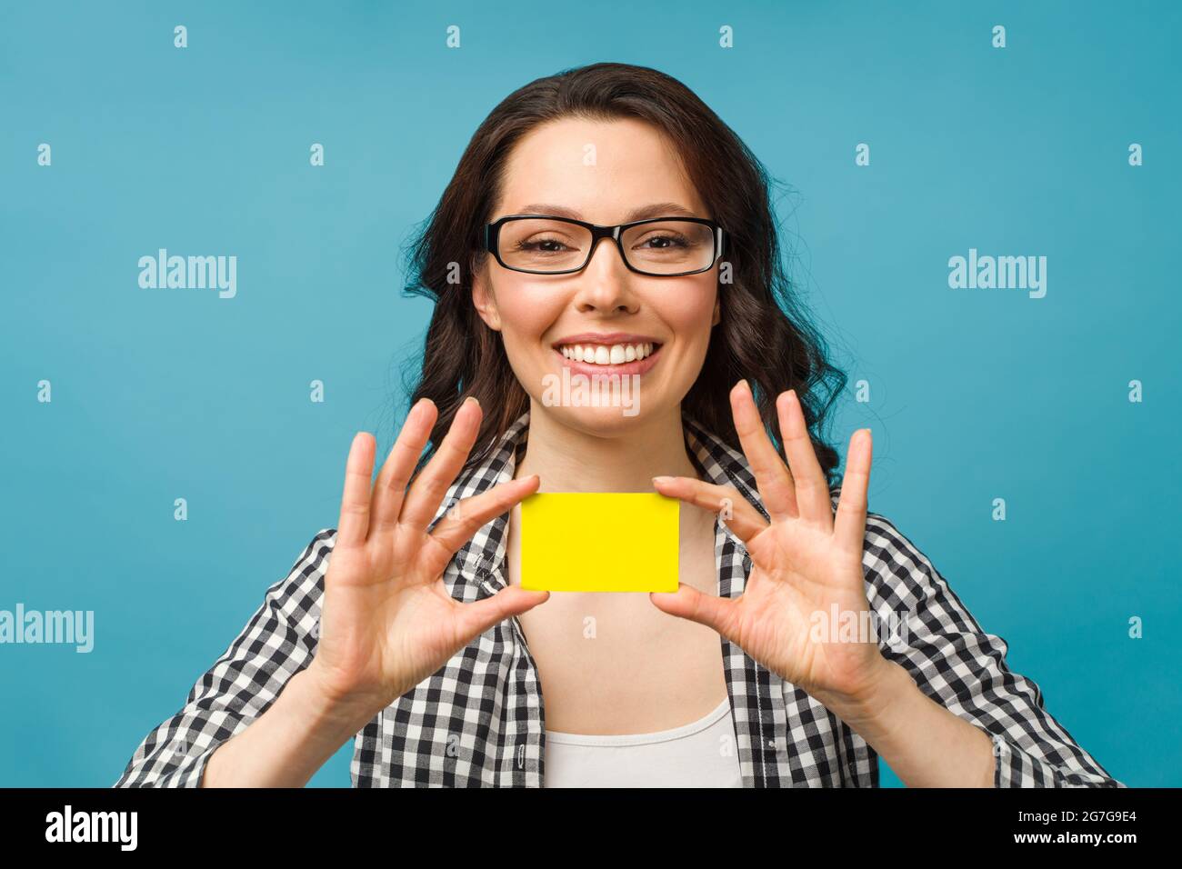 Jolie femme souriante dans une chemise habillée et des lunettes, montrant la carte de crédit à la main pour le concept de société financière et sans cashless, posant sur fond bleu Banque D'Images