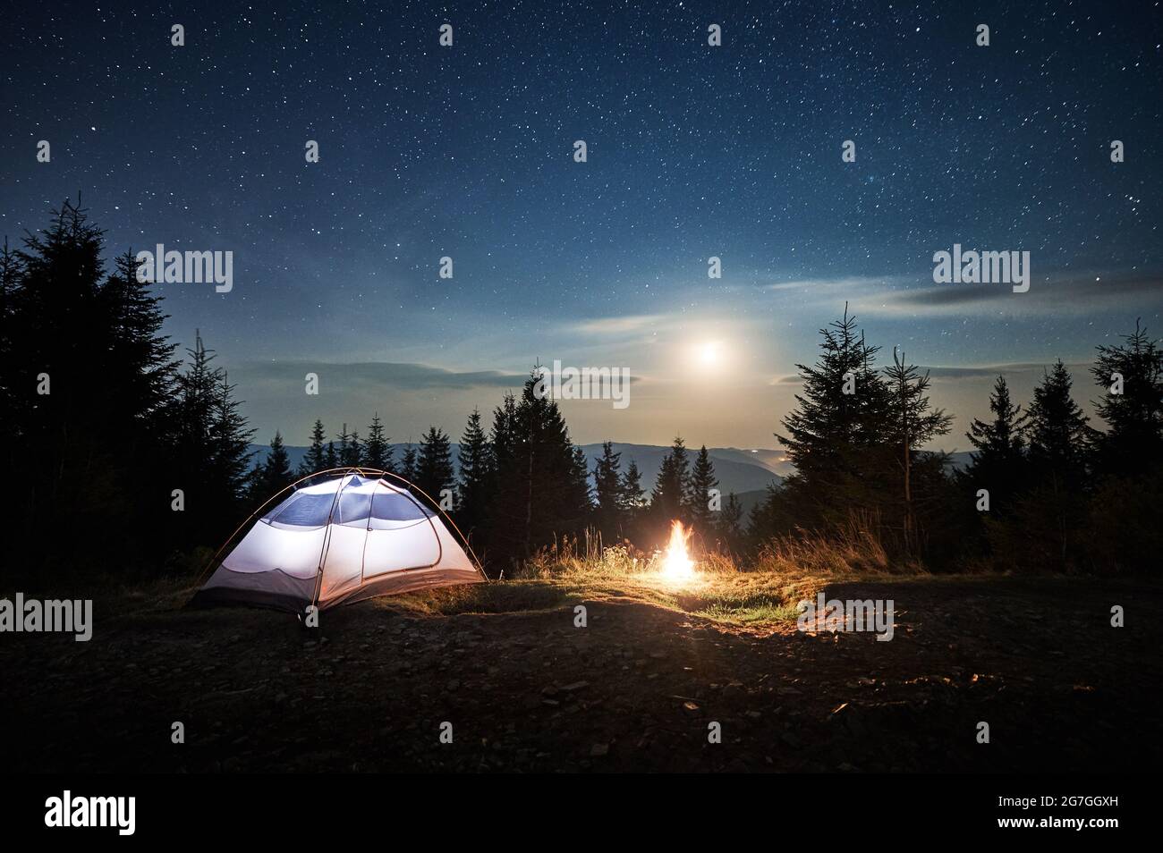 Tente touristique blanche située sur une colline de montagne près d'un feu de camp en feu de forêt. Moonlight dans le ciel étoilé magique nuit camping dans les montagnes. Concept de camping de nuit. Banque D'Images