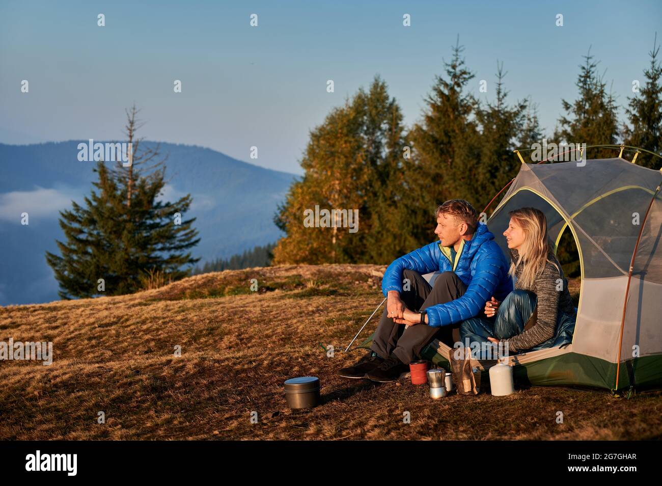 Vue latérale de couple de voyageurs mariés se reposant dans une tente sur un pré de montagne pendant la randonnée d'automne sur la toile de fond des arbres et de la colline de montagne. Matin de soleil dans le camping. Banque D'Images