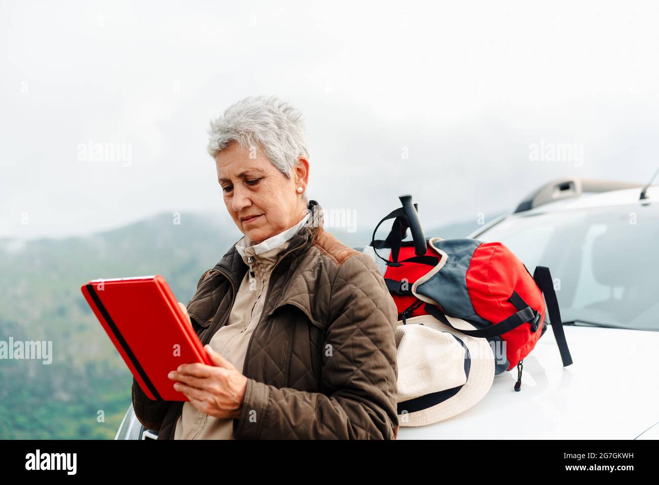 Femme voyageur âgée aux cheveux gris courts penchée sur la voiture et parcourant une tablette lors d'un voyage en campagne Banque D'Images