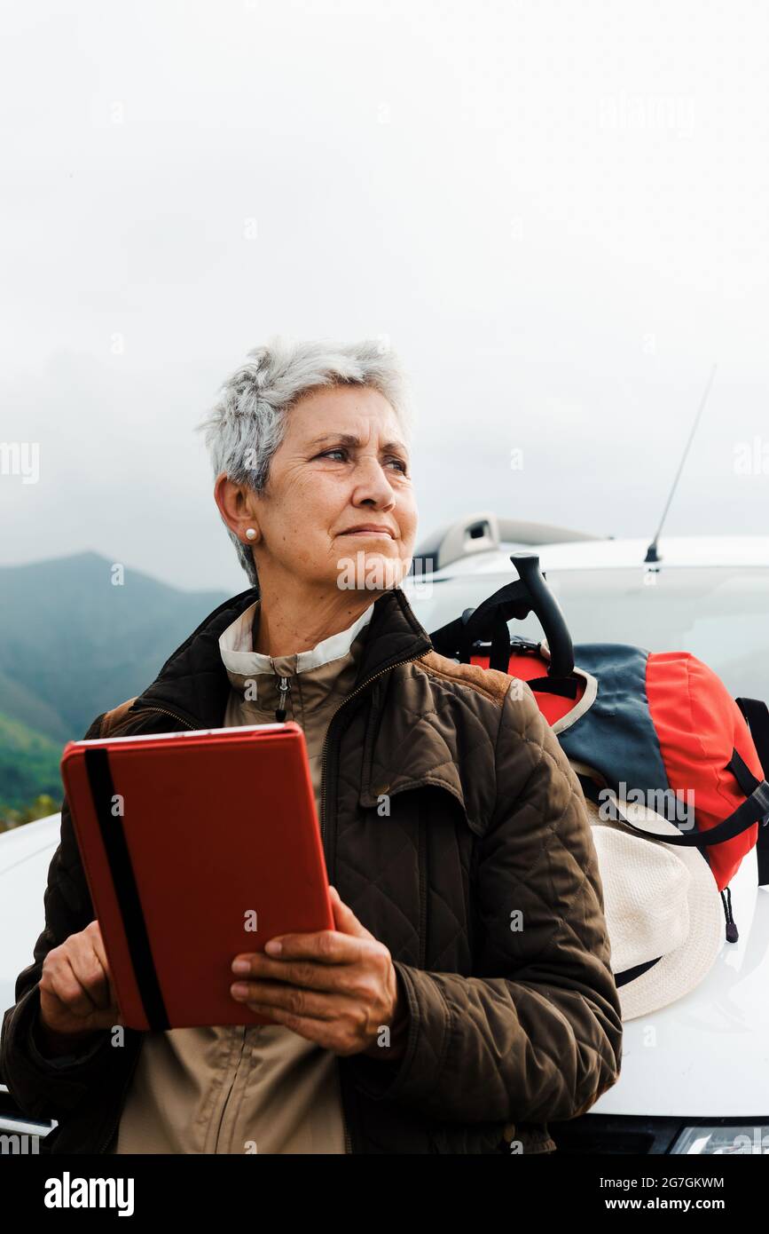 Voyageur âgé avec de courts cheveux gris penchée sur la voiture et parcourant une tablette pendant un voyage en campagne et regardant loin Banque D'Images