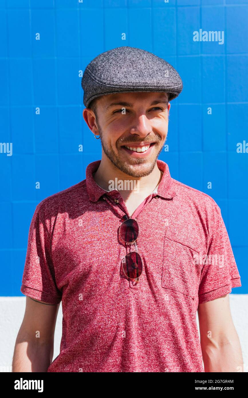Joyeux jeune homme barbu dans un style décontracté et casquette profiter de la journée d'été près du mur bleu sur la rue urbaine Banque D'Images
