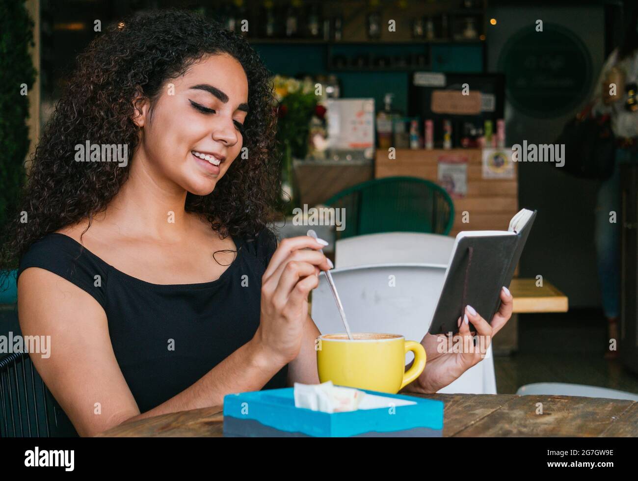Jeune femme hispanique souriante aux cheveux maurires, en train de se reposer à table avec une tasse de café sur la terrasse du café Banque D'Images