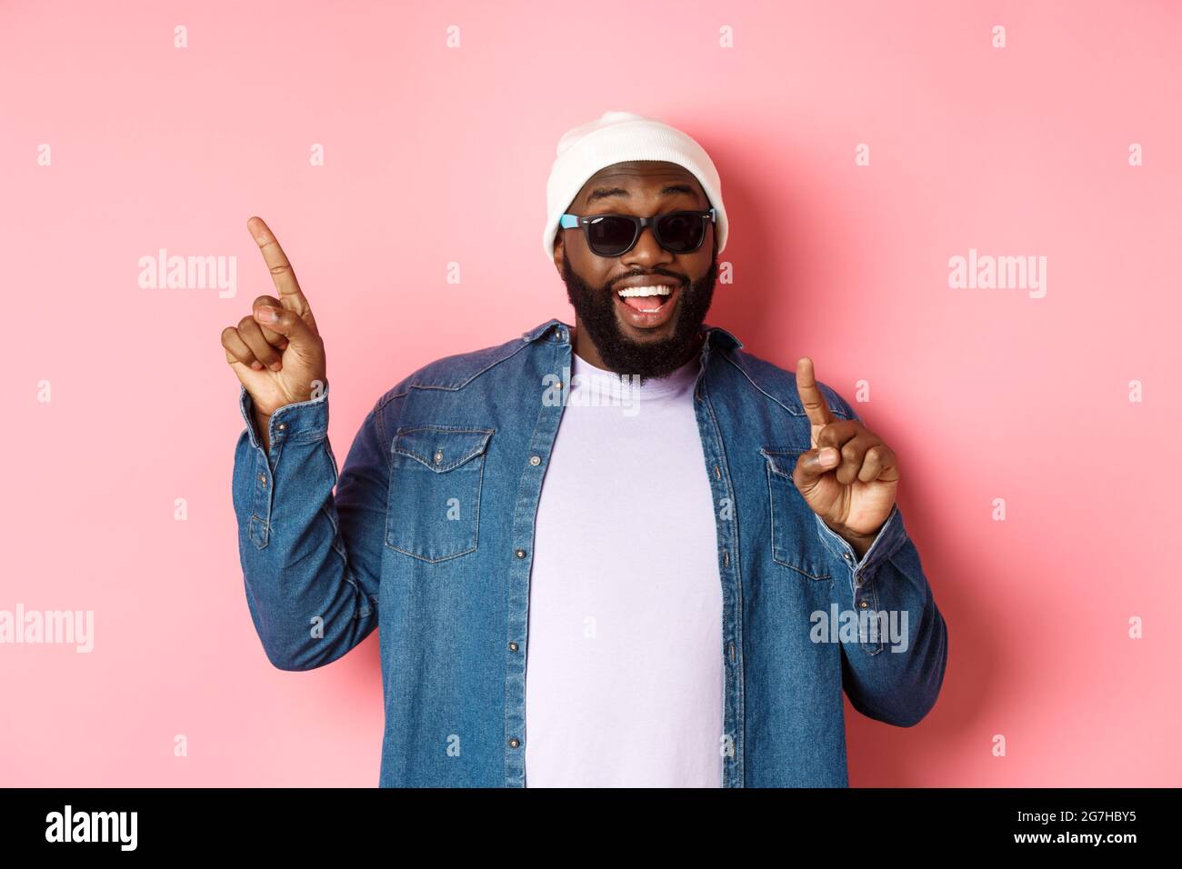 Joyeux homme afro-américain à barbe montrant l'annonce, pointant les doigts dans le coin supérieur gauche et souriant, debout dans un bonnet et des lunettes de soleil sur rose Banque D'Images