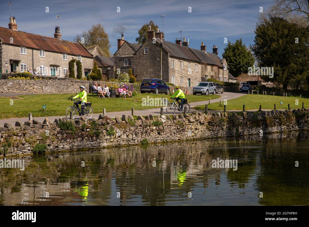 Royaume-Uni, Angleterre, Derbyshire, Tissington, The Green, cyclistes âgés en vestes haute viz, de l'autre côté de l'étang du village Banque D'Images