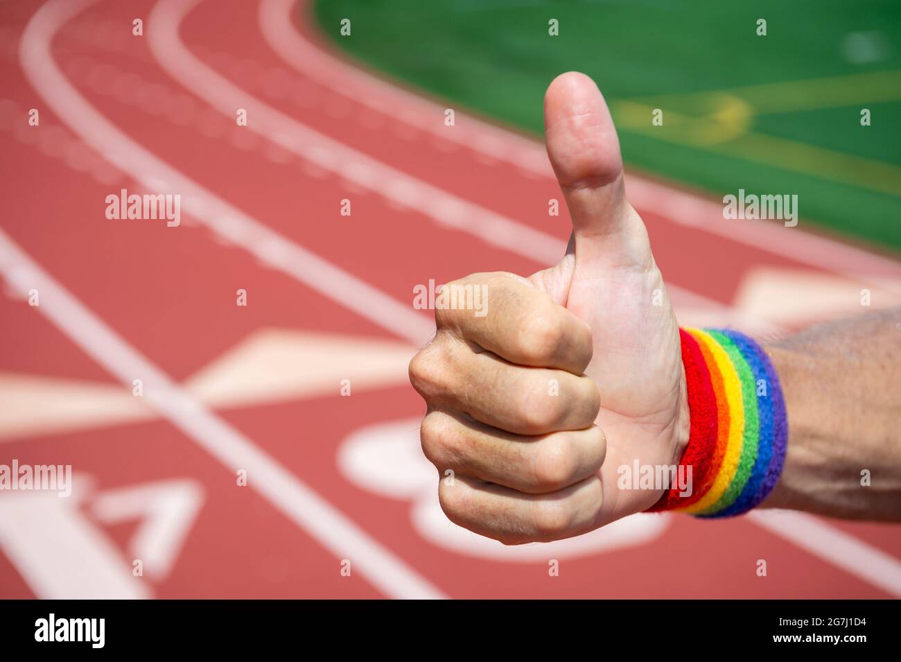 L'athlète gay avec le bracelet de fierté arc-en-ciel donne le pouce vers le haut de geste devant un fond de piste de sport Banque D'Images