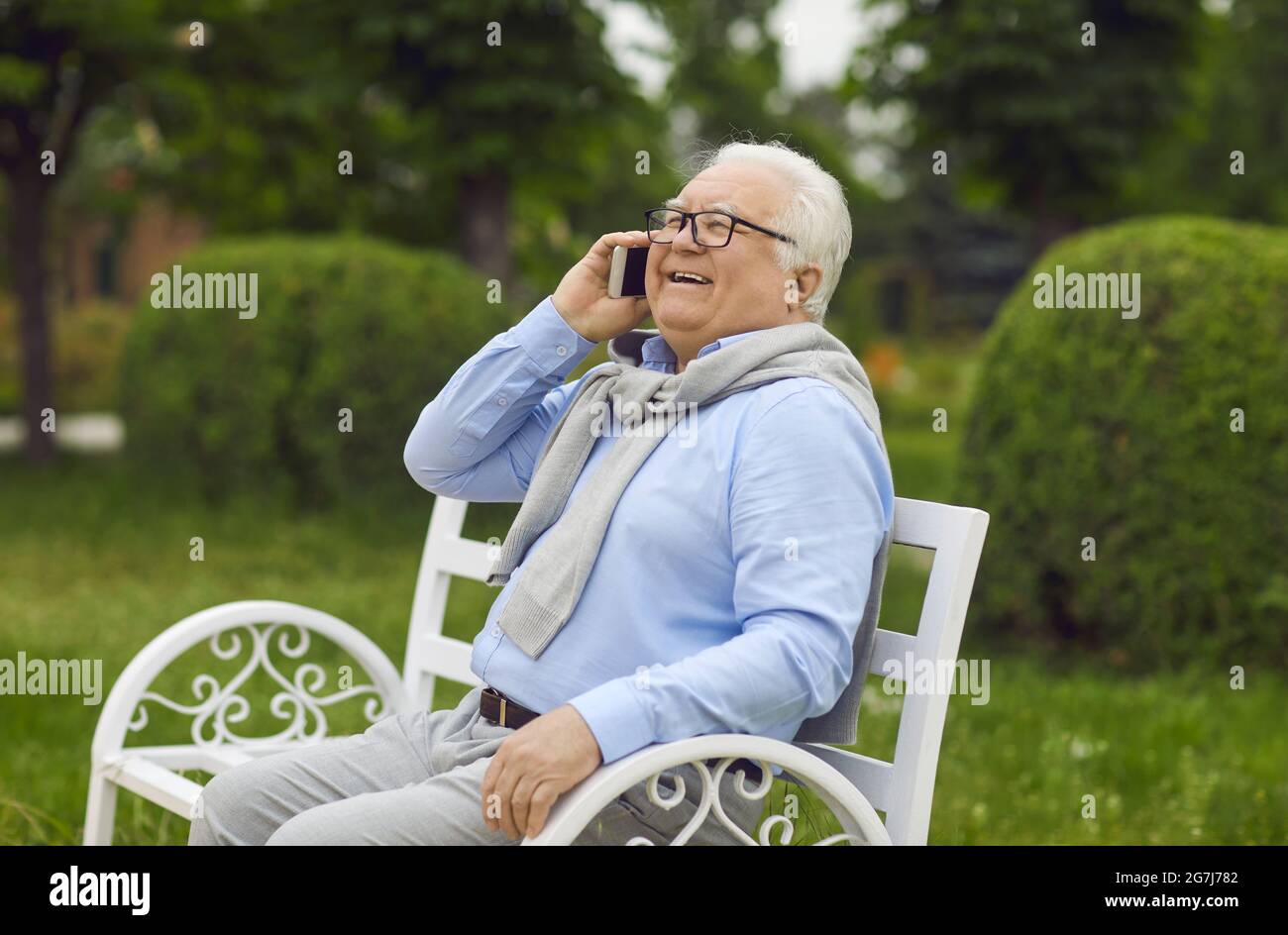 Joyeux homme senior parlant sur téléphone portable tout en étant assis sur le banc dans le parc vert d'été Banque D'Images