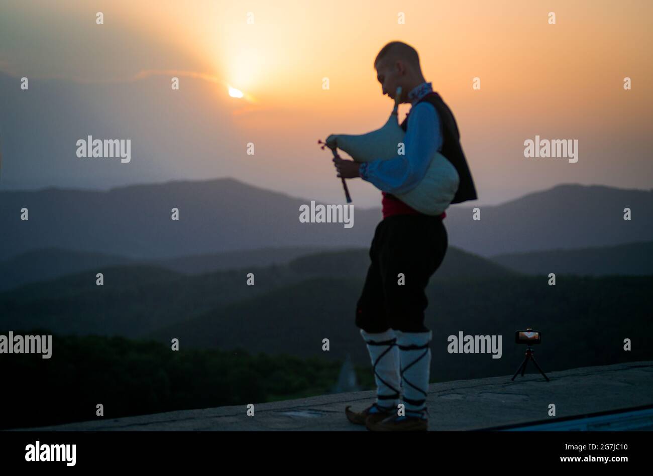 Un jeune joueur de cornemuse en costume folklorique joue sur un pic de montagne quand le soleil se lève, le matin 01 juillet 2021, Shipka, Bulgarie Banque D'Images