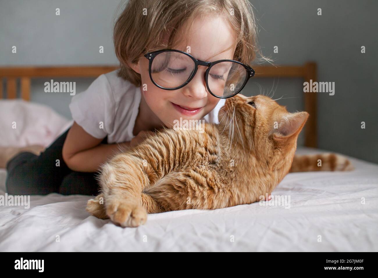 une petite fille avec des verres se trouve sur le lit et embrasse un gros chat de gingembre. Banque D'Images