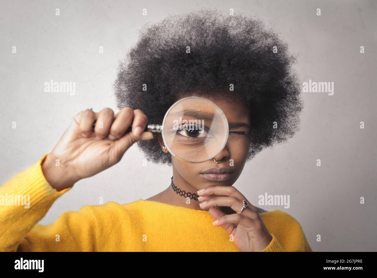 Belle jeune femme noire regardant à travers une loupe contre un mur blanc Banque D'Images