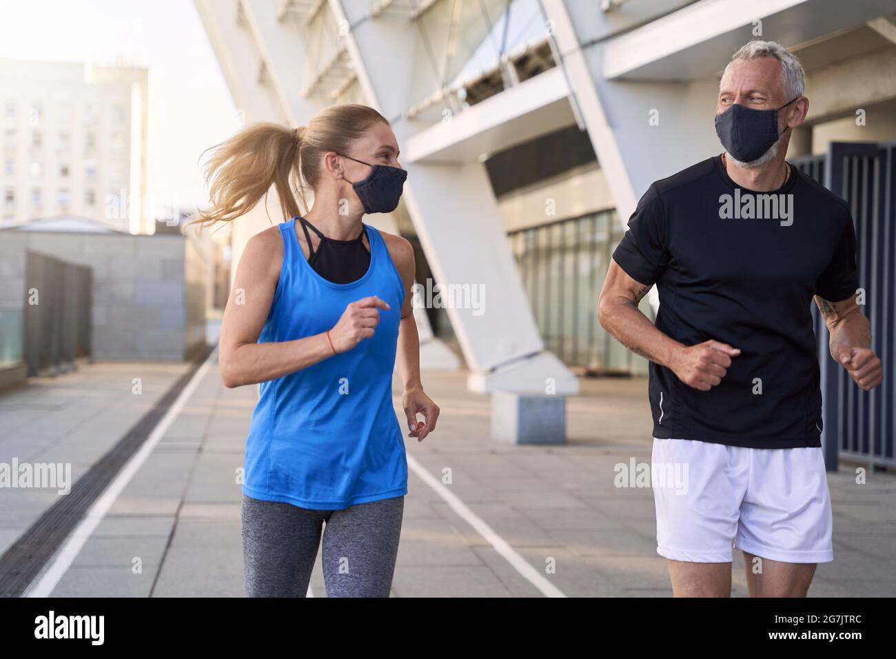 Couple d'âge moyen motivé portant des masques protecteurs se regardant les uns les autres tout en courant ensemble en milieu urbain pendant le coronavirus Banque D'Images