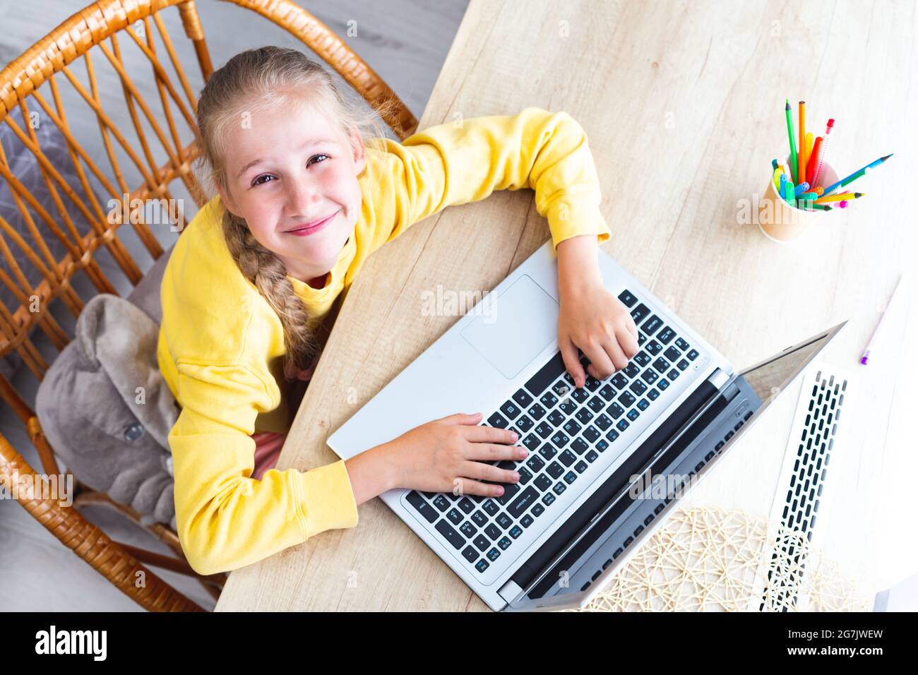 Caucasien belle fille de 10-11 ans s'assoit dans une chaise en osier à un bureau en bois clair, les mains sur le clavier d'ordinateur portable, regardant la caméra, sourires Banque D'Images