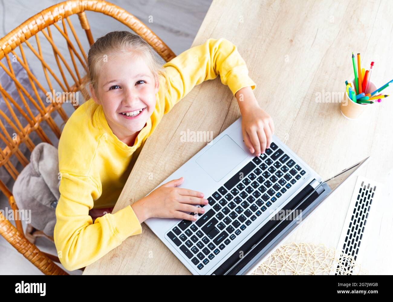 Caucasienne heureuse fille de 10-11 ans est assise dans une chaise en osier à un bureau en bois clair, les mains sur un clavier d'ordinateur portable, regardant la caméra, vue de dessus Banque D'Images