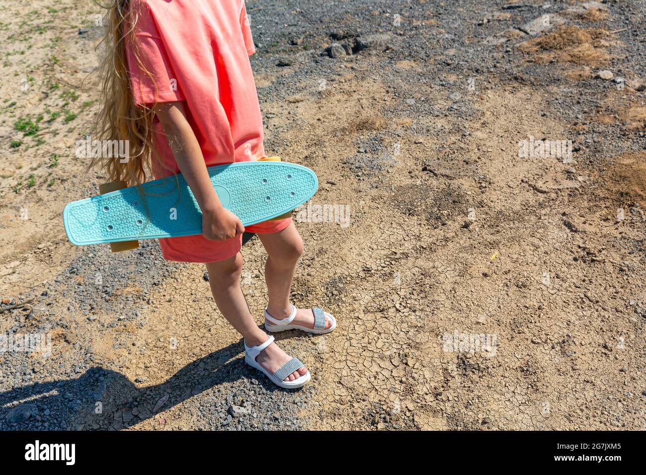 Une jeune fille adolescente mince avec un skateboard à la main dans un costume tendance d'été avec des cheveux blonds flatteur sur un fond de terre craquée sur un Banque D'Images