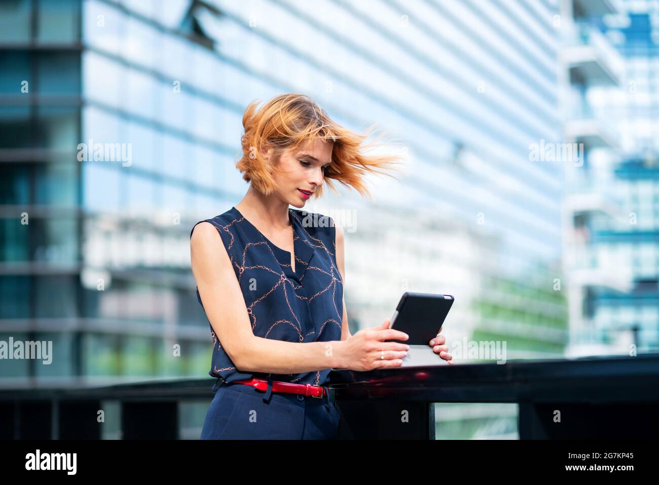 Belle femme élégante debout regardant des médias ou lisant en ligne sur sa tablette à l'extérieur sur un pont urbain avec fond de gratte-ciel et des cheveux de Blui Banque D'Images