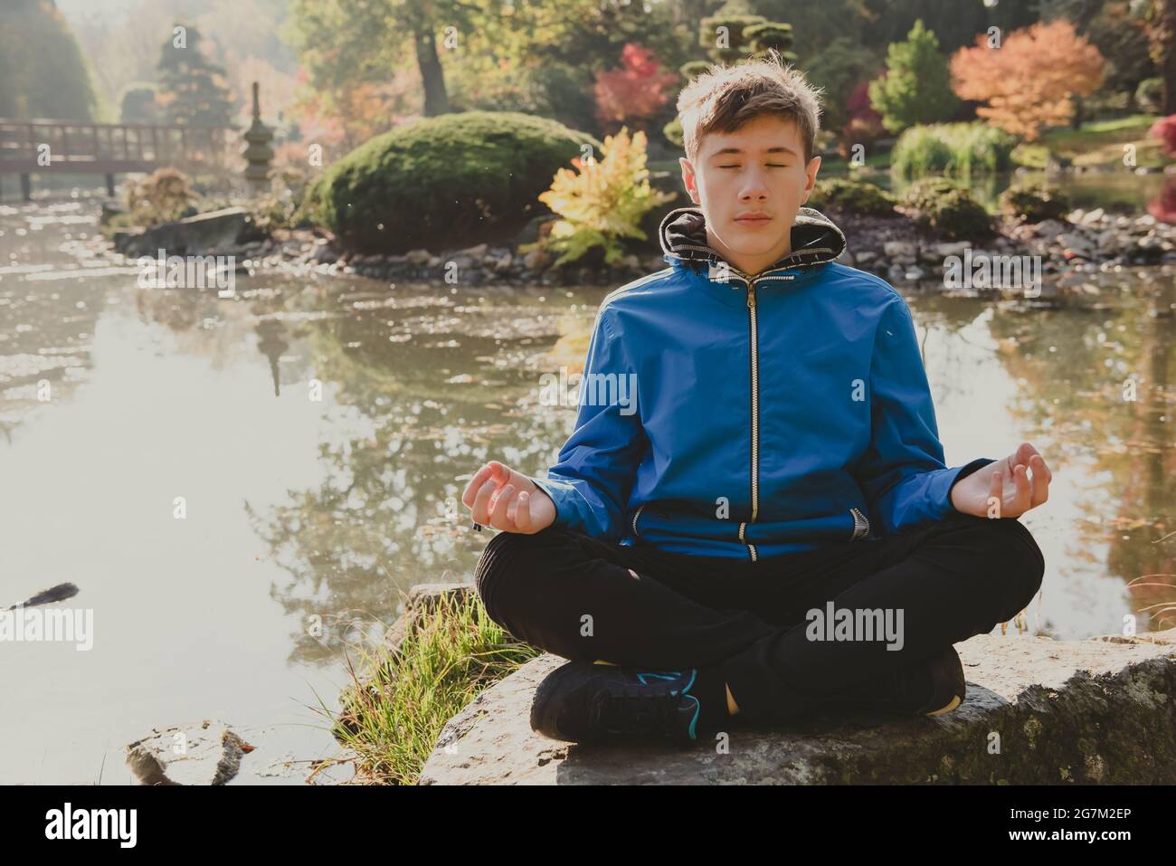 Jeune garçon méditant et relaxant dans un jardin japonais de parc de ville. Jeune garçon dans une position de lotus méditant, méditation en plein air sur une pierre sur un étang s. Banque D'Images