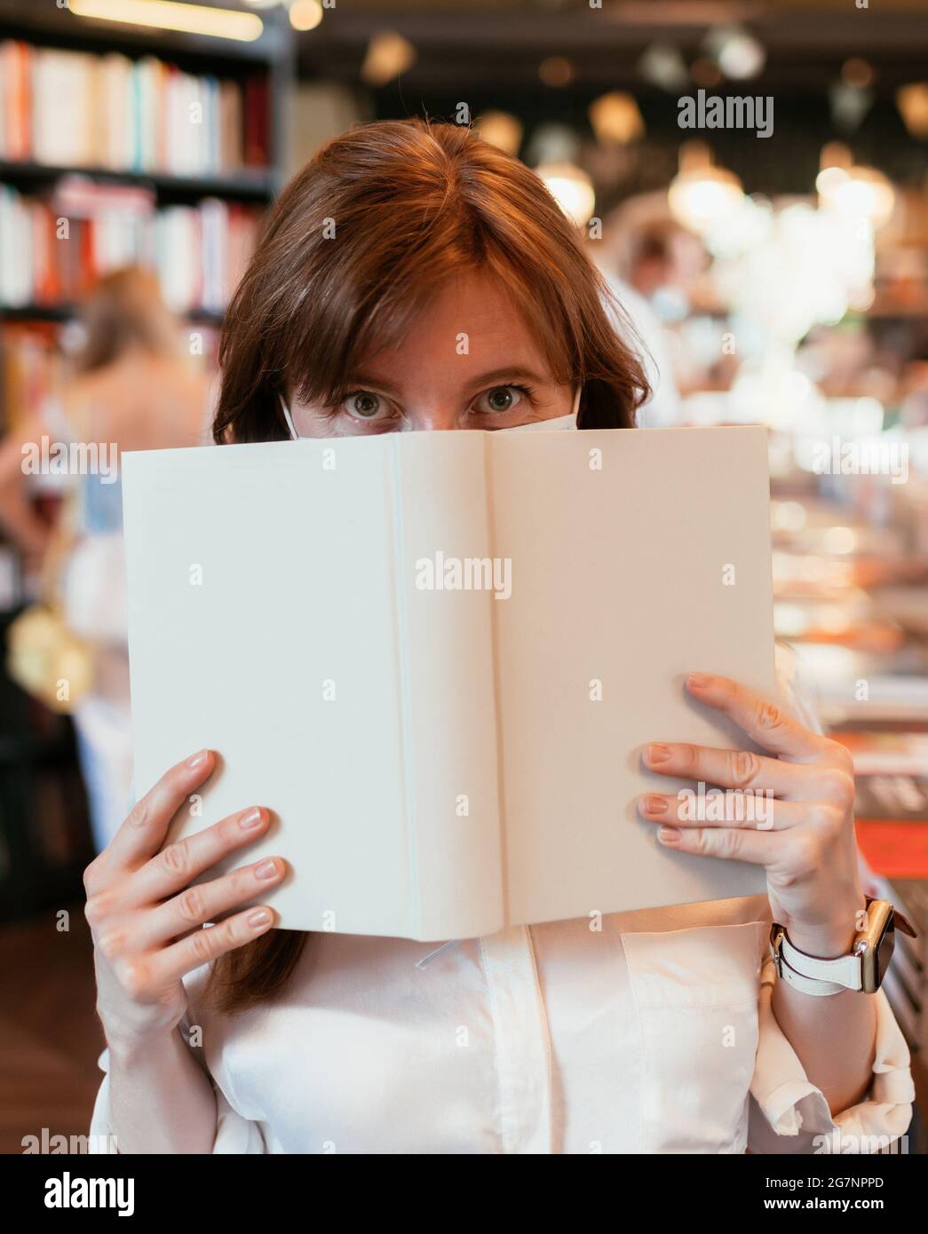 Fille dans un masque médical dans la bibliothèque couvre la moitié de son visage avec un livre. Banque D'Images