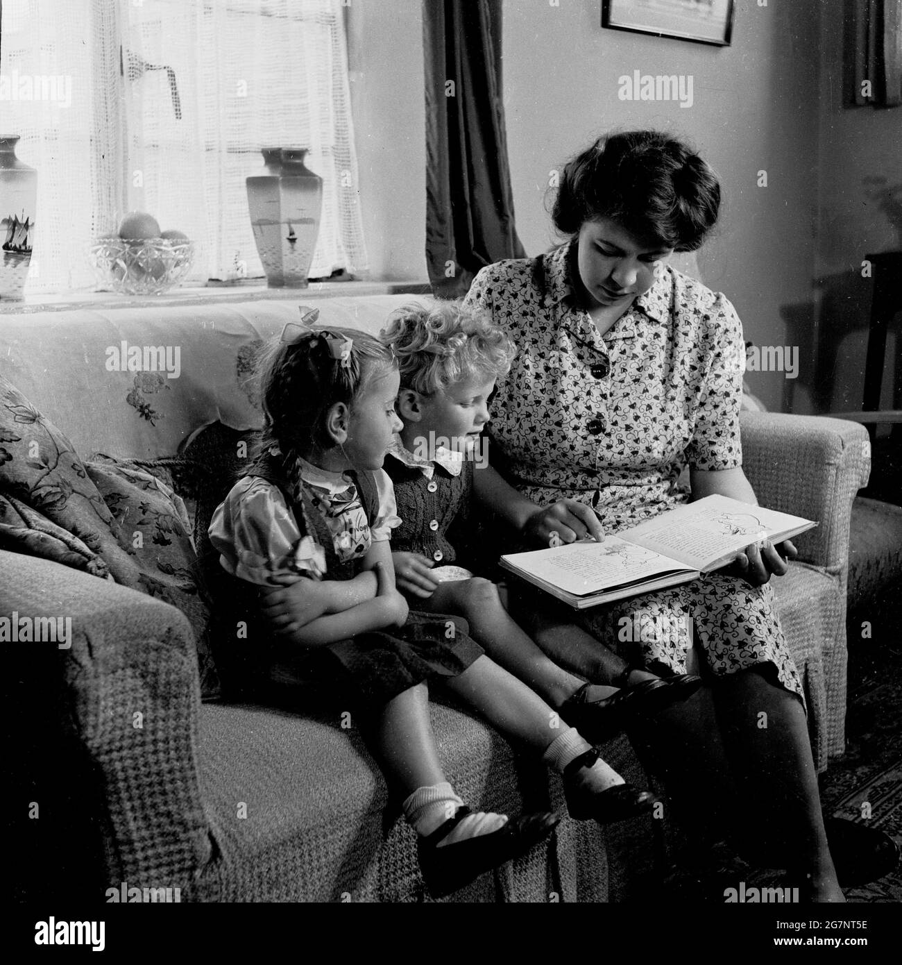 Années 1950, historique, une mère assise sur un canapé avec ses deux jeunes enfants les lisant une histoire d'un livre, Angleterre, Royaume-Uni. Banque D'Images