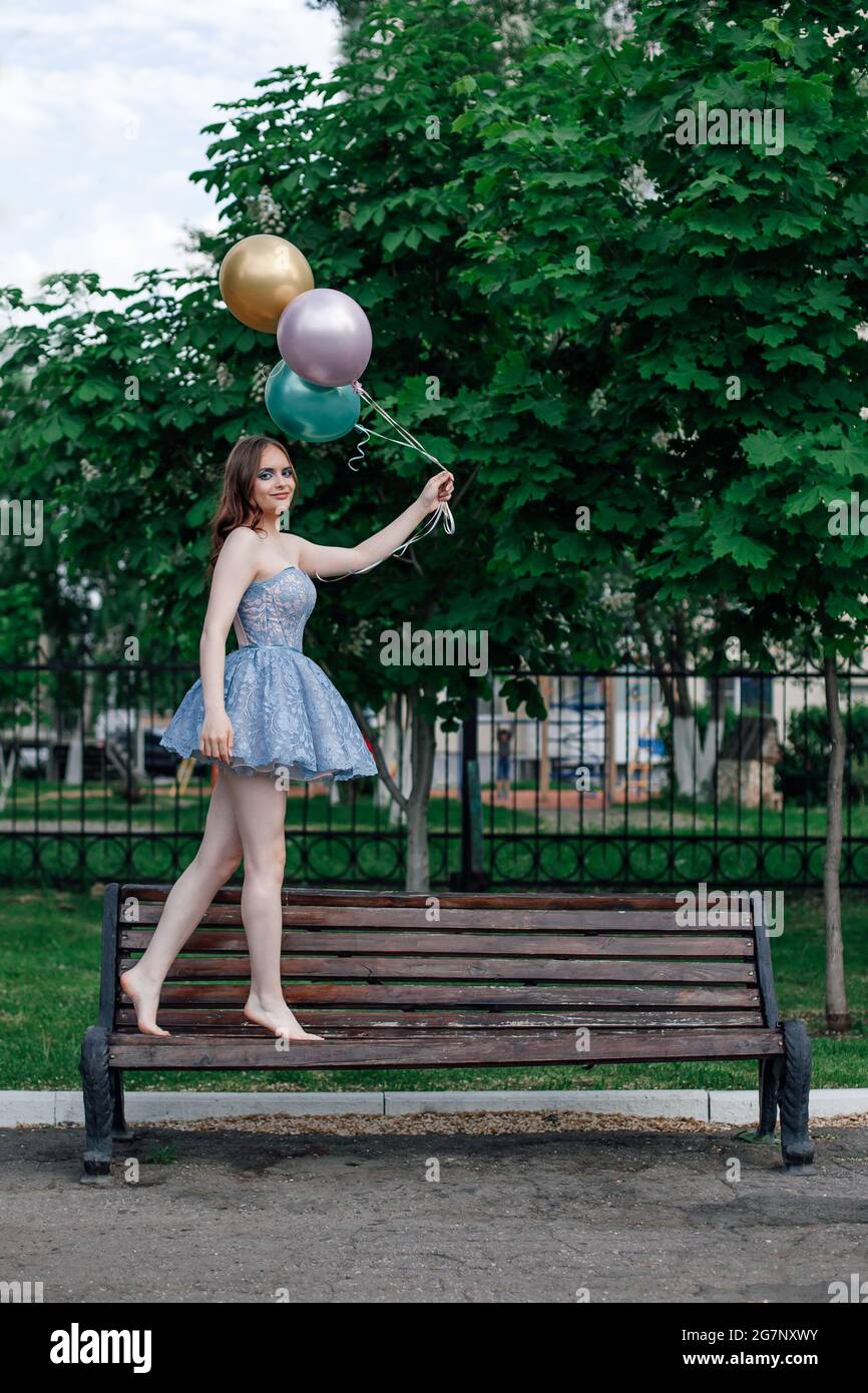 une jeune femme heureuse, vêtue d'un short corset bleu, marche pieds nus  sur un banc et tient des ballons volants, une fille romantique sur une date  avec des ballons Photo Stock -