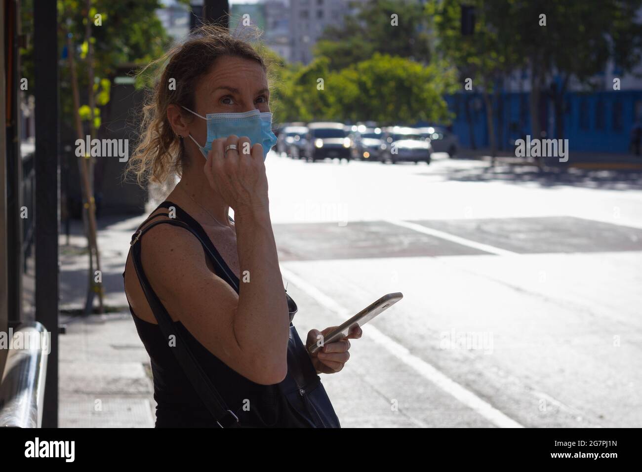 Femme d'âge moyen avec couverture faciale attendant à la gare routière tenant le téléphone portable. Femme solitaire avec masque dans la rue. Concept de distanciation sociale Banque D'Images