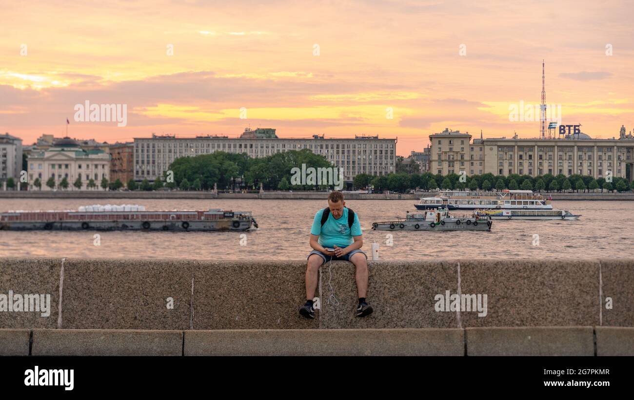 Un homme qui vérifie son smartphone tout en étant assis sur un remblai de granit avec vue panoramique au coucher du soleil sur la rivière Neva avec des bateaux la nuit blanche, Saint-Pétersbourg, Rus Banque D'Images