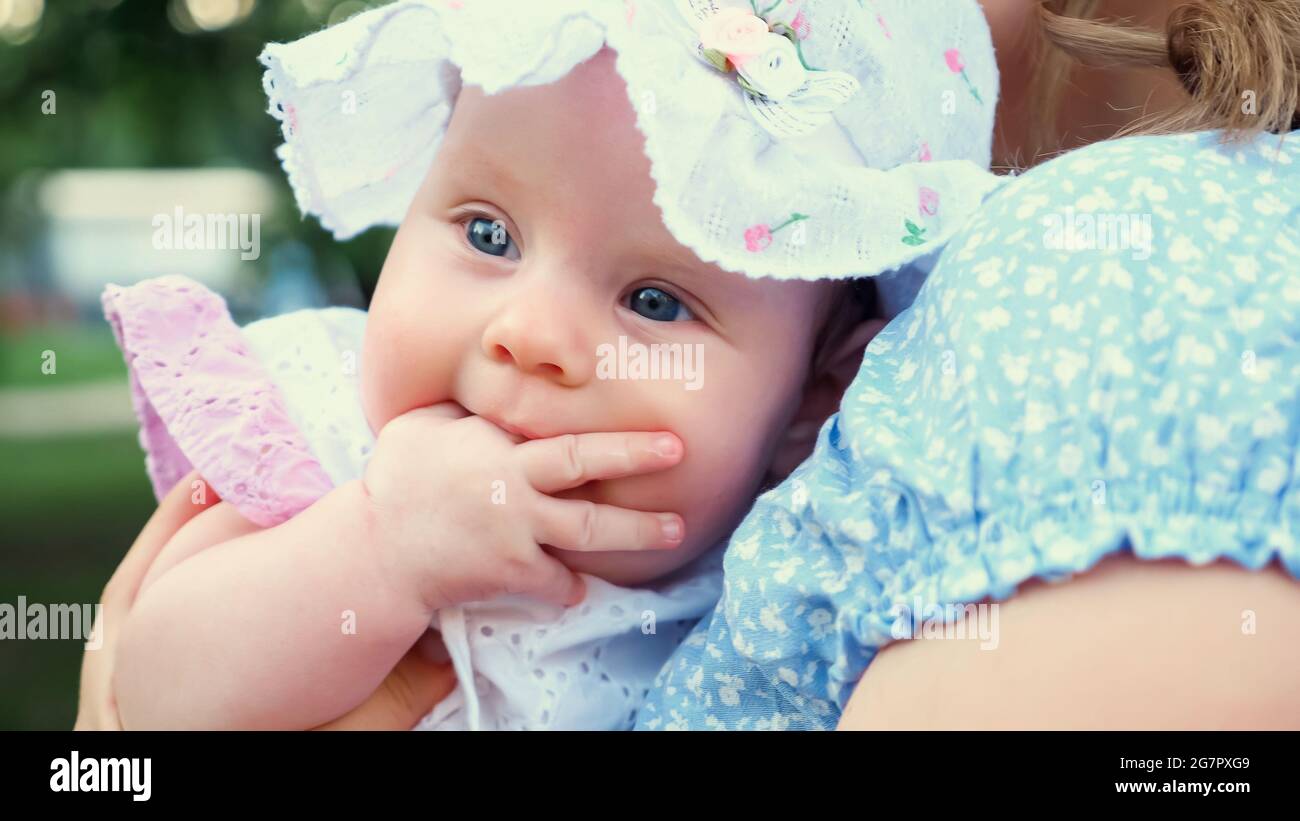 Mignon bébé fille avec les yeux bleus au panama regarde autour et suce le  doigt assis dans les bras blonds de mère sous la lumière du soleil de  printemps vue étroite Photo