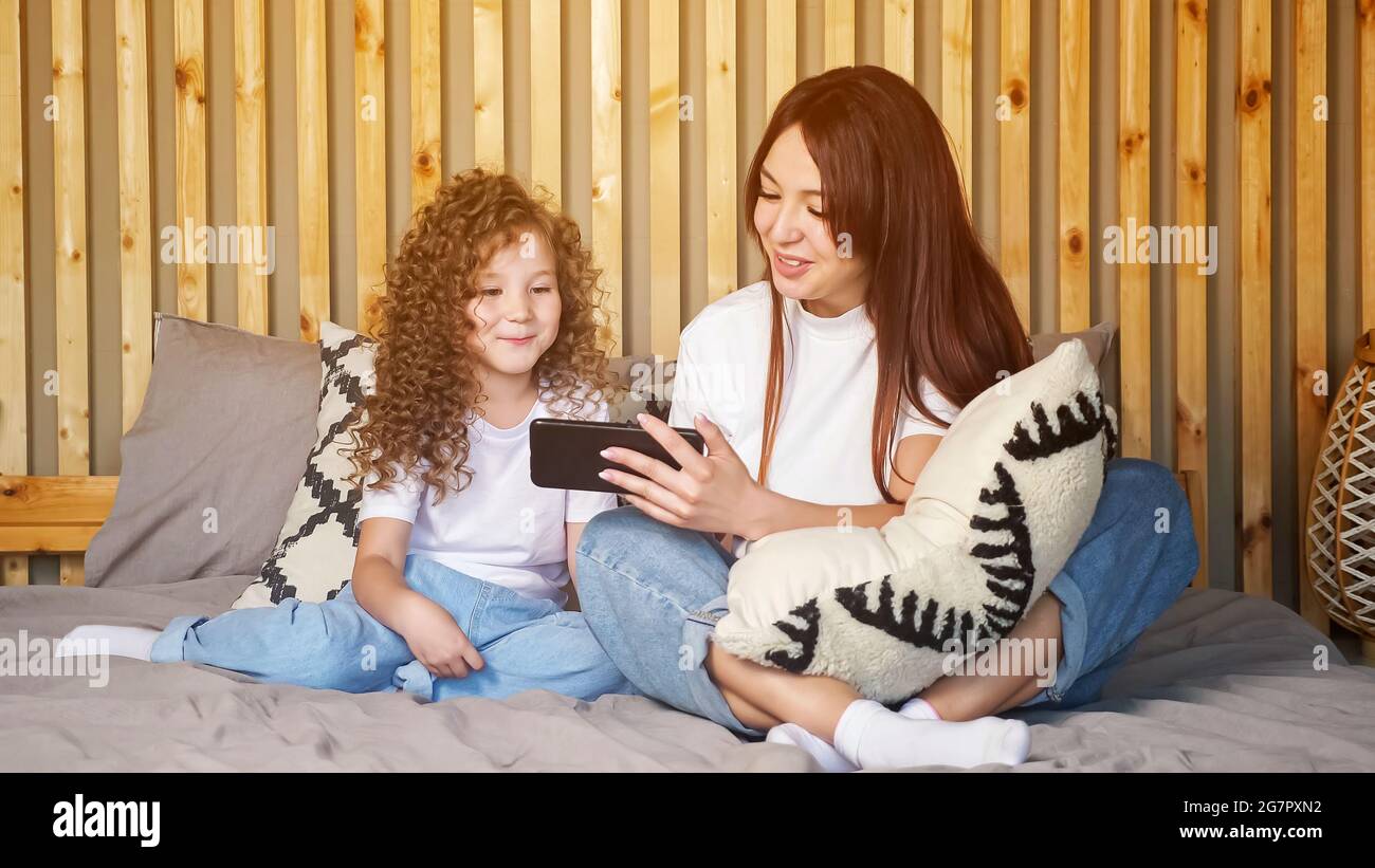 Brunette mère et enfant fille parler avec le père sur appel vidéo en agitant les mains et les baisers soufflants, sur grand lit gris contre le mur en bois brun à la maison Banque D'Images