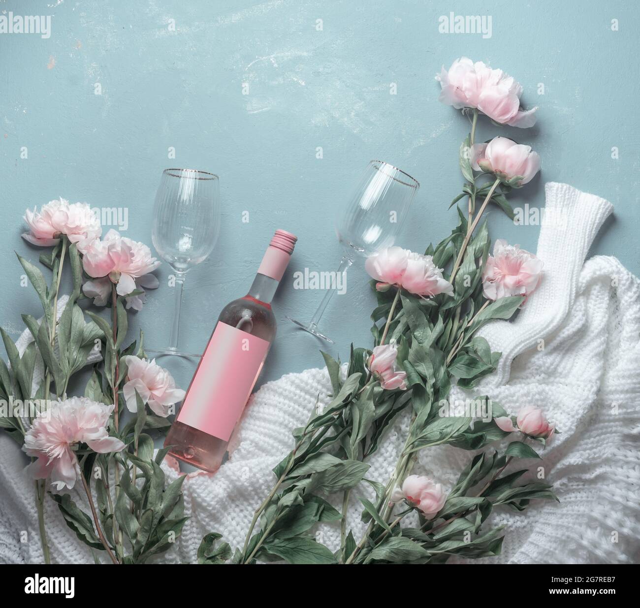 Bouteille de vin rose avec des lunettes de soleil sur fond bleu avec chandail blanc tricoté et de nombreuses fleurs de péonies roses pâles. Vue de dessus. Beauté Banque D'Images