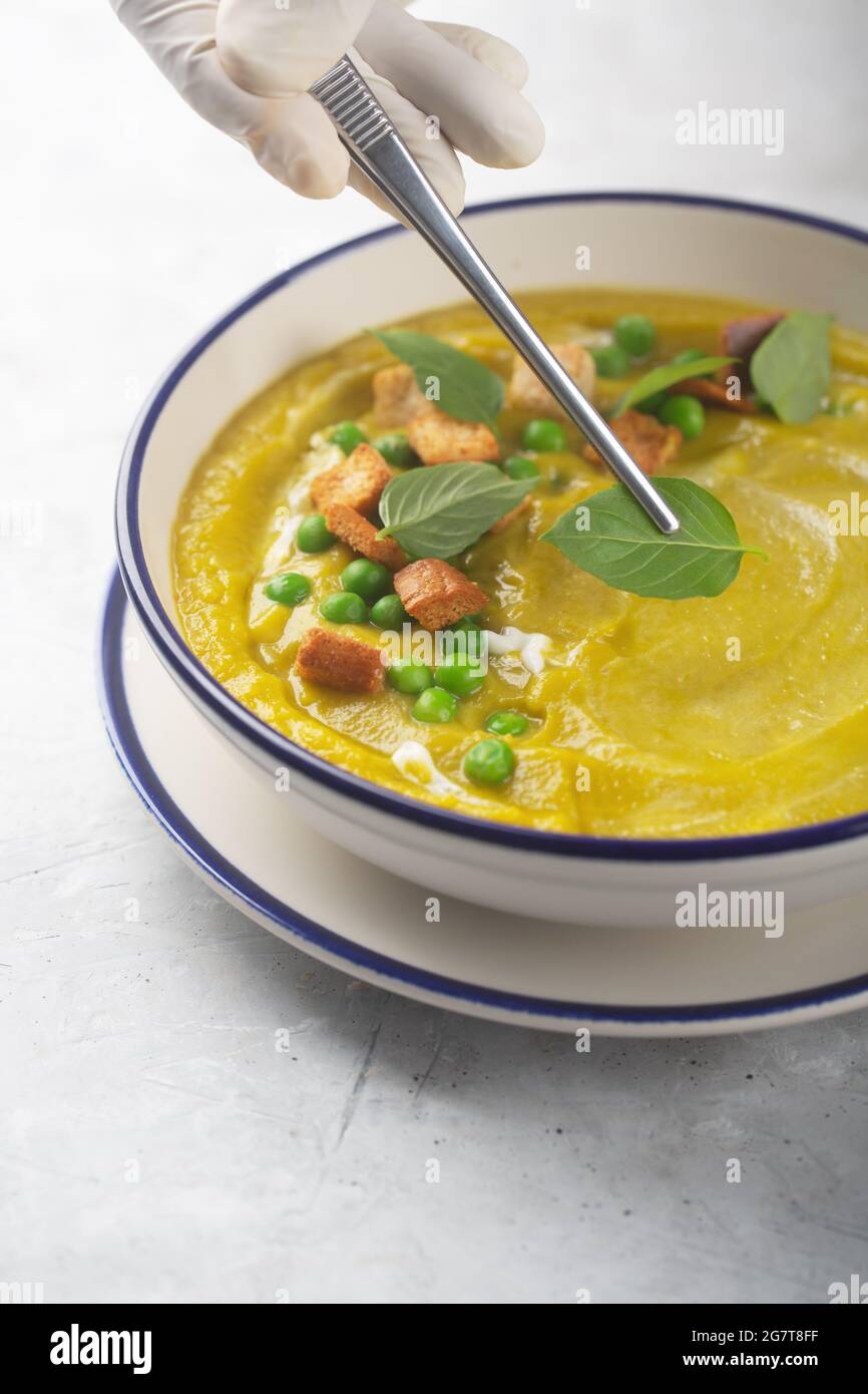 Un chef en gants décorera une soupe de légumes biologiques en purée avec des craquelins, des pois verts et des herbes aux feuilles de basilic . Bonne nutrition.nourriture de placage Banque D'Images