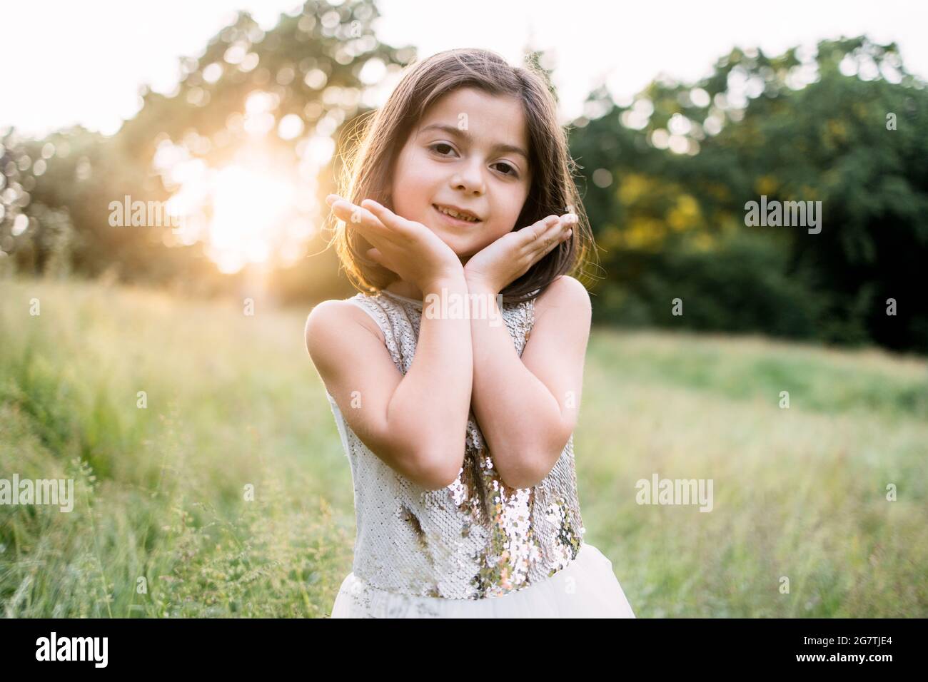 Portrait d'une jolie petite fille aux cheveux bruns courts, en tenant ses mains sur le menton et les joues, en posant au jardin vert avec un beau lever de soleil derrière. Concept de bonheur et d'enfance. Banque D'Images