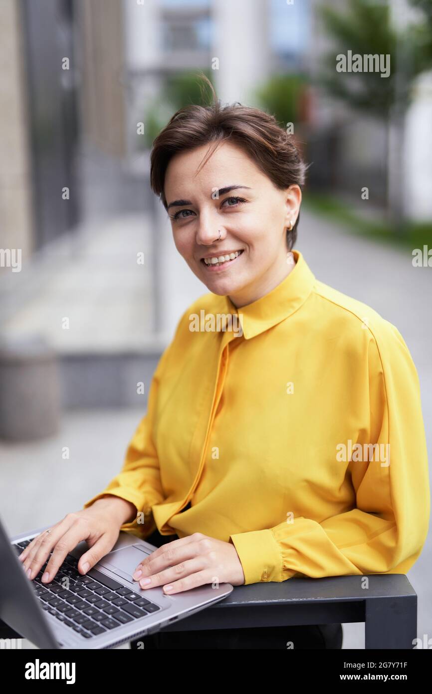 Gros plan portrait d'une femme d'affaires heureuse et qualifiée, jeune femme d'affaires des années 30, qui séjourne dans la rue avec un ordinateur portable et regarde l'appareil photo. Femme entrepreneur travaillant à l'extérieur du bâtiment de bureau. Image de haute qualité Banque D'Images