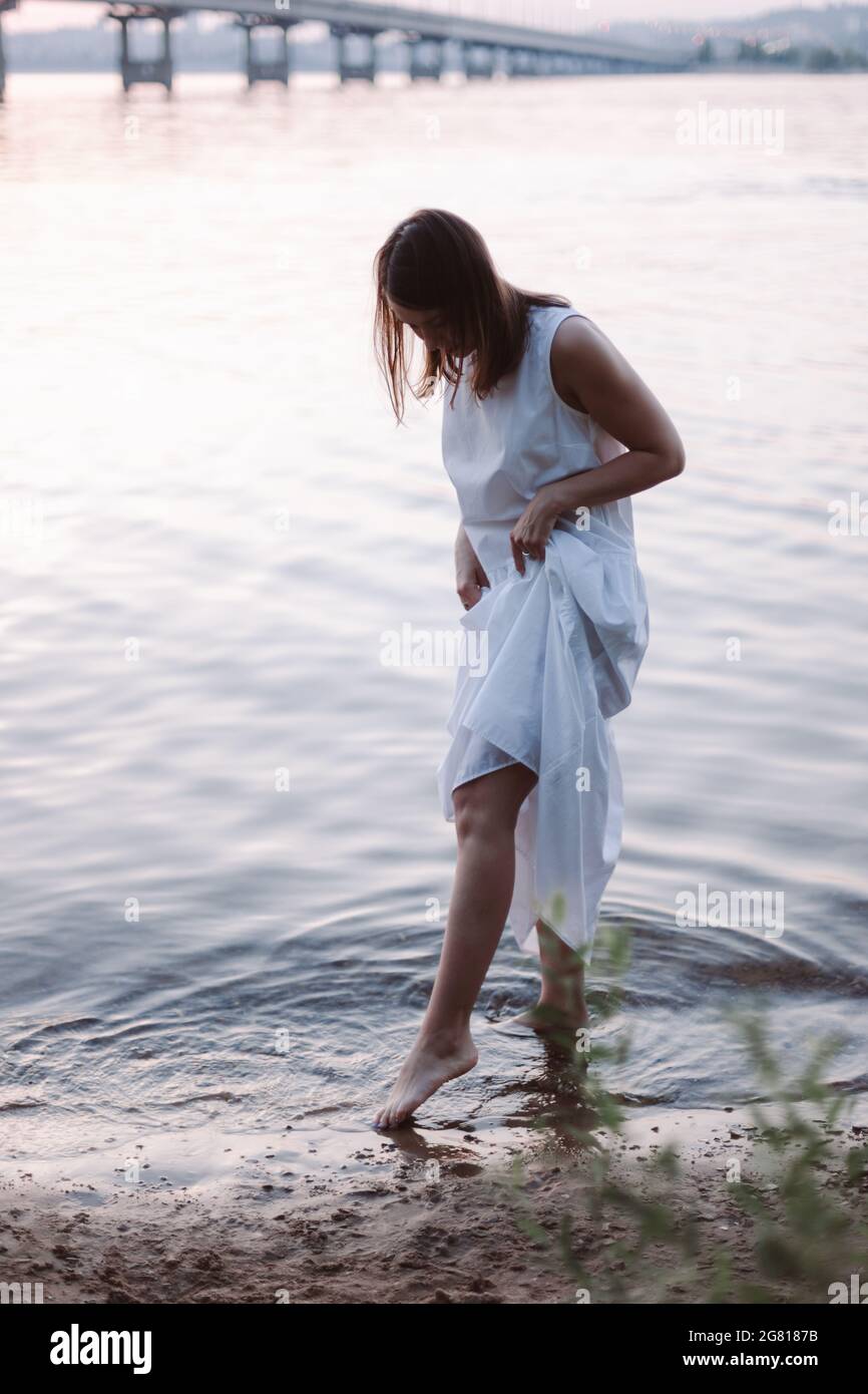 une jeune femme mince vêtue d'une robe blanche d'été, levant l'ourlet de sa  robe, se lance dans l'eau sur la plage de sable de la rivière après le  coucher du soleil Photo