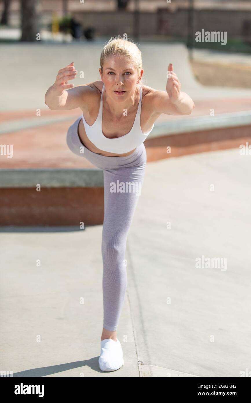 La femme blonde atractive en bonne santé et en forme montre la concentration et l'équilibre face à la ligne droite sur une jambe au skate Park. Banque D'Images